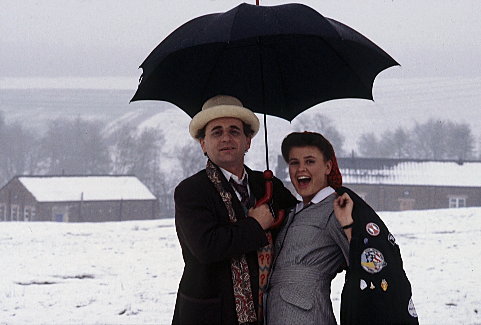 Sophie Aldred as Ace and Sylvester McCoy as the Seventh Doctor standing under an umbrella